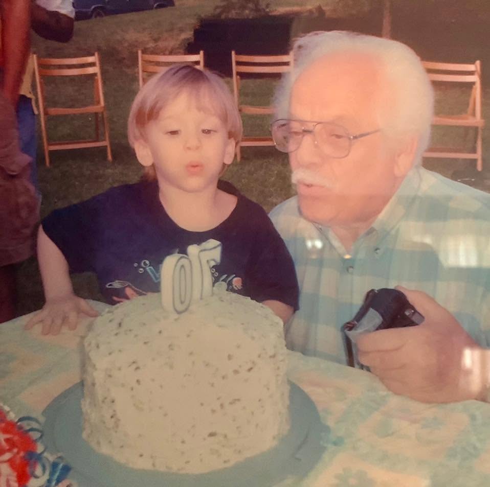 Nathaniel as a child, blowing out the candles on a birthday cake, with his grandfather leaning in close beside him, camera in hand